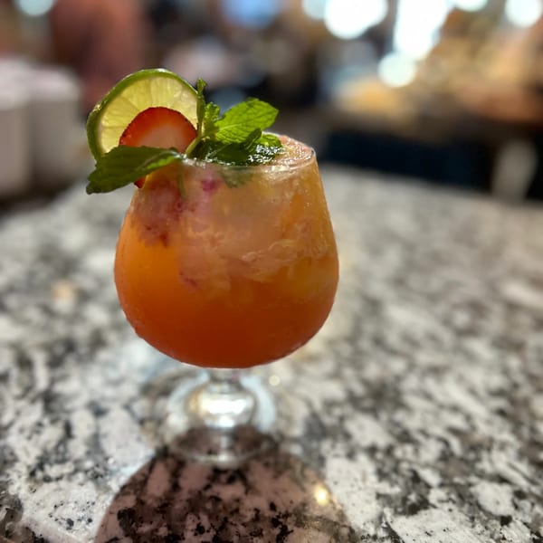 A vibrant orange cocktail garnished with lime, strawberry, and mint sits on a speckled countertop.